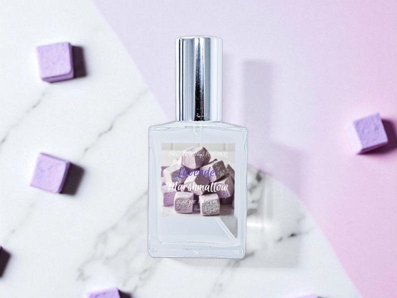 May include: A clear glass perfume bottle with a silver cap, featuring a label with the words "Love de Marshmallow" over a graphic of purple marshmallow cubes. Scattered around the bottle are more purple marshmallow cubes. The background is a mix of white marble and light purple.