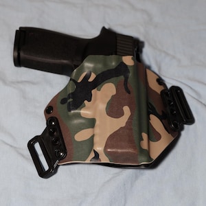 May include: A black handgun is placed in a holster with a camouflage design. The holster is patterned with green, brown, and beige. It has black belt loops and fittings. The firearm is partially visible.
