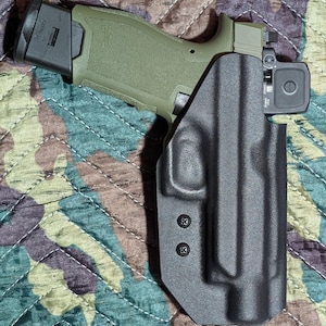 May include: An olive green handgun with a black magazine and a black holster. The handgun has a red dot sight. The holster is black and secured with two black screws. The background is a camouflage pattern.