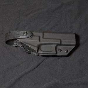 May include: A black polymer holster with a belt loop. The holster has a contoured design with screw points and a textured surface. The image is set against a dark background.