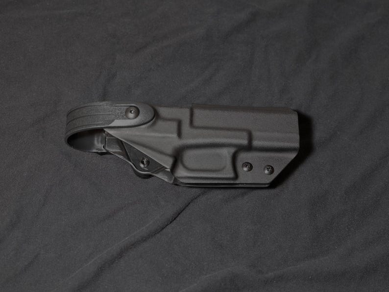 May include: A black polymer holster with a belt loop. The holster has a contoured design with screw points and a textured surface. The image is set against a dark background.