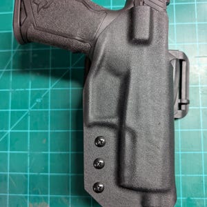 Puede incluir: Una funda de polímero negro con un arma de fuego en su interior. La funda tiene una superficie texturizada y se asegura con tres tornillos negros. La empuñadura del arma es visible, con un patrón texturizado y un logotipo.