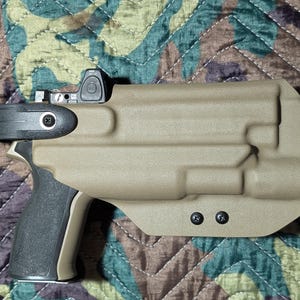 May include: Tan colored pistol holster with a black grip and a mounted optic. The holster has a molded design with two black screws. The background is a camouflage pattern.