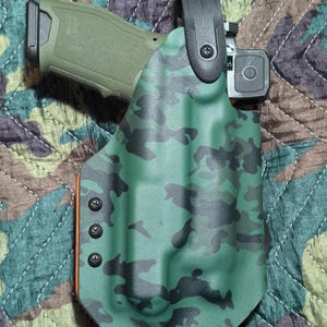 Könnte beinhalten: Eine grüne und schwarze Tarnmusterholster mit einem schwarzen Gürtelclip. Die Holster hält eine grüne Handfeuerwaffe mit einem schwarzen Schlitten und einem Rotpunktvisier. Die Holster ist mit schwarzen Schrauben befestigt und hat einen orangefarbenen Akzent.