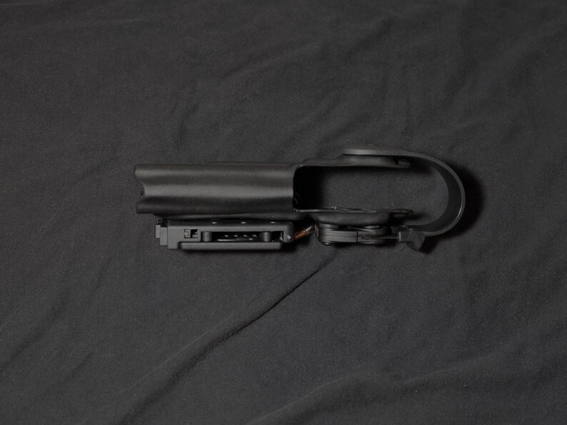 May include: A black plastic holster, designed for a firearm. It has a belt loop and a trigger guard. The object is set against a dark background.