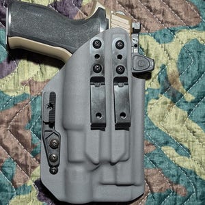Op de afbeelding: Een grijze holster met zwarte hardware houdt een vuurwapen vast met een zwarte grip en een tan slide. De holster heeft twee riemclips en een retentie-apparaat. De achtergrond is een camouflagepatroon.