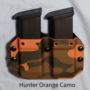 Może przedstawiać: Podwójny uchwyt na magazynki w kolorze Hunter Orange Camo. Uchwyt posiada dwa czarne magazynki, zabezpieczone wzorem kamuflażu z pomarańczowymi i brązowymi odcieniami. Czarne śruby mocują uchwyt.