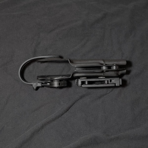 May include: A black plastic holster with a curved belt clip and a rectangular attachment. The holster is designed to hold a firearm. The image is set against a dark background.