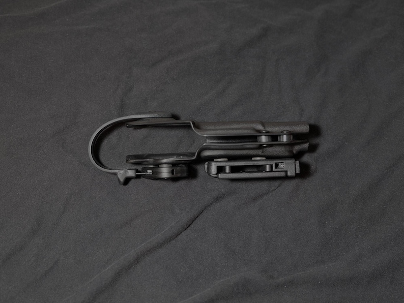 May include: A black plastic holster with a curved belt clip and a rectangular attachment. The holster is designed to hold a firearm. The image is set against a dark background.