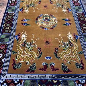 Puede incluir: Una alfombra rectangular de color marrón dorado con un diseño intrincado que presenta cuatro dragones dorados y un motivo central de dragón. La alfombra tiene un borde azul oscuro con patrones geométricos y una franja blanca.