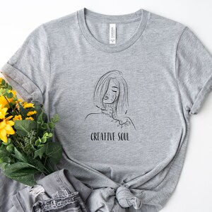Pu&ograve; includere: Maglietta grigia con un disegno a linea nera di una donna con fiori sulla spalla e il testo "CREATIVE SOUL".