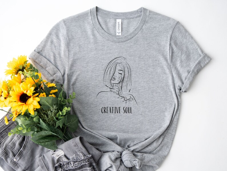Pu&ograve; includere: Maglietta grigia con un disegno a linea nera di una donna con fiori sulla spalla e il testo "CREATIVE SOUL".