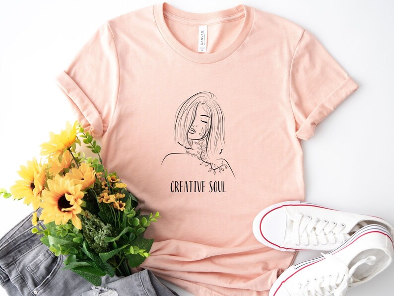 Pu&ograve; includere: Una maglietta color pesca con un disegno a linea nera di una donna con fiori sul viso e il testo "CREATIVE SOUL".