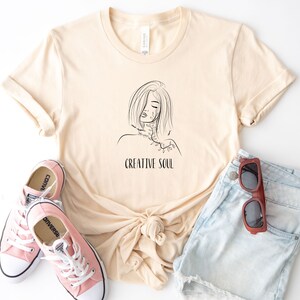 Pu&ograve; includere: Una maglietta beige con un disegno a linea nera di una donna con un motivo floreale sul collo e il testo "CREATIVE SOUL" sotto il disegno.
