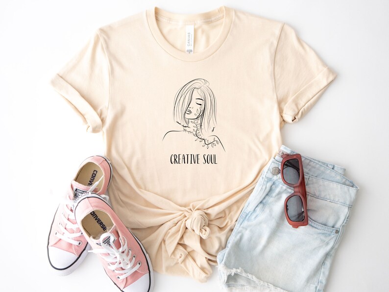 Pu&ograve; includere: Una maglietta beige con un disegno a linea nera di una donna con un motivo floreale sul collo e il testo "CREATIVE SOUL" sotto il disegno.