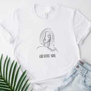 Pu&ograve; includere: Maglietta bianca con un disegno a linea nera di una donna con fiori intorno al collo e il testo "CREATIVE SOUL" sotto l'immagine.