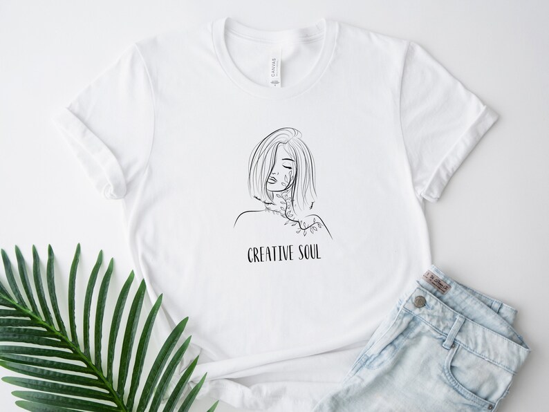 Pu&ograve; includere: Maglietta bianca con un disegno a linea nera di una donna con fiori intorno al collo e il testo "CREATIVE SOUL" sotto l'immagine.
