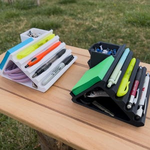 May include: Two desk organizers, one white and one black, sit on a wooden surface. The white organizer holds pens, highlighters, and a bundle of purple cords. The black organizer contains pens, sticky notes, and other office supplies.