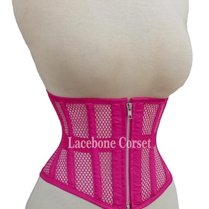 May include: A hot pink corset with a mesh and vertical panel design. The corset has a front zipper closure and the text "Lacebone Corset" is visible. The corset is displayed on a mannequin.
