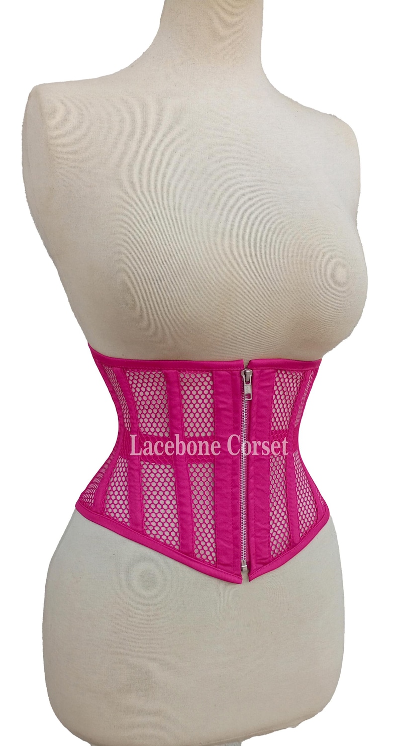 May include: A hot pink corset with a mesh and vertical panel design. The corset has a front zipper closure and the text "Lacebone Corset" is visible. The corset is displayed on a mannequin.
