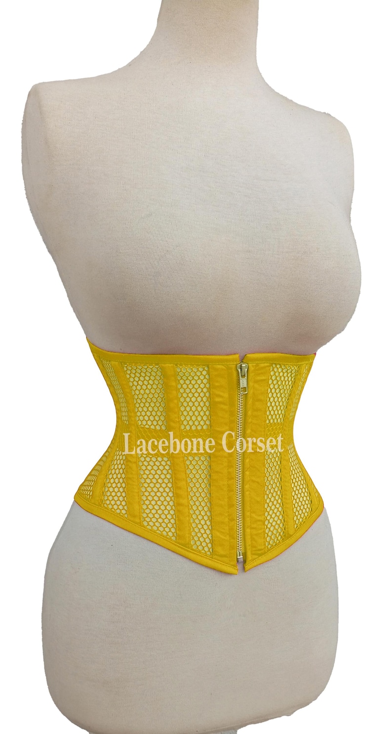 May include: A bright yellow corset with a mesh and vertical panel design. The corset has a front zipper closure and is displayed on a mannequin. The text "Lacebone Corset" is visible on the front.