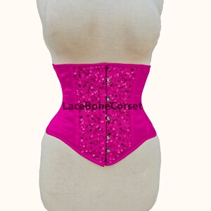 May include: A hot pink satin corset with a decorative design of sequins and silver buttons. The corset has a lace-up back and is a popular choice for special occasions.