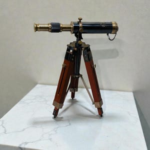 Może przedstawiać: Miniaturowy, zabytkowy teleskop z mosiężnymi i czarnymi akcentami, zamontowany na drewnianym statywie. Korpus teleskopu jest czarny z mosiężnymi okuciami, a statyw ma czerwonobrązowe nogi. Całkowita wysokość około 15 cm.