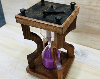 May include: A wooden hourglass with a dark finish and a square top. The top has a dark metal plate with decorative elements and four black knobs. The hourglass contains purple sand. The wooden frame has a unique, open design.