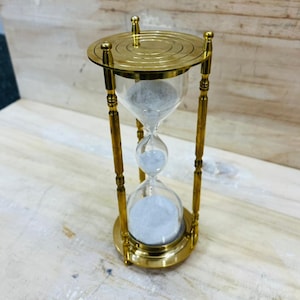 Puede incluir: Un reloj de arena de latón con un centro de vidrio transparente lleno de arena blanca. El reloj de arena tiene una base y una parte superior circulares, conectadas por cuatro pilares de latón. El diseño es vintage y decorativo.