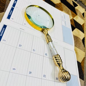 May include: A gold-coloured magnifying glass with a mother-of-pearl and gold handle lies on an open calendar. The magnifying glass has a round lens and a decorative handle. The calendar shows the days of the week and the month of July.
