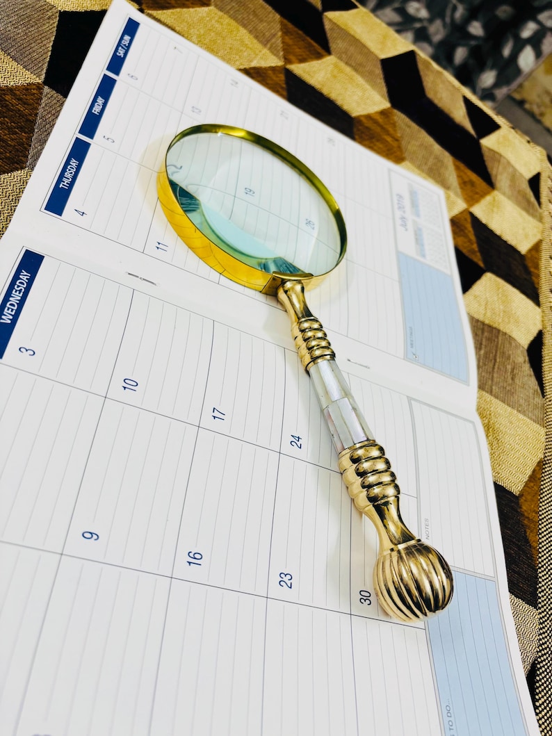 May include: A gold-coloured magnifying glass with a mother-of-pearl and gold handle lies on an open calendar. The magnifying glass has a round lens and a decorative handle. The calendar shows the days of the week and the month of July.