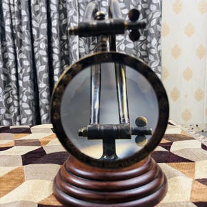 May include: A brass magnifying glass with a wooden base. The magnifying glass is on a stand and has a round lens. The stand is made of dark wood and has a decorative design.