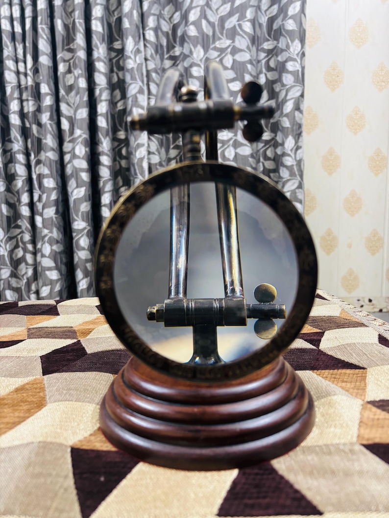 May include: A brass magnifying glass with a wooden base. The magnifying glass is on a stand and has a round lens. The stand is made of dark wood and has a decorative design.