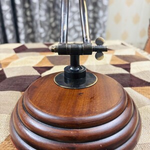 May include: A wooden base with three tiers, topped with a black metal base and a brass adjustable arm. The arm has a brass knob and a brass wing nut.