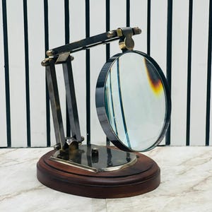 May include: A magnifying glass mounted on an adjustable metal arm with a dark wood oval base. The circular lens is framed in dark metal and attached to a multi-jointed stand that allows for positioning. The base is polished dark wood.