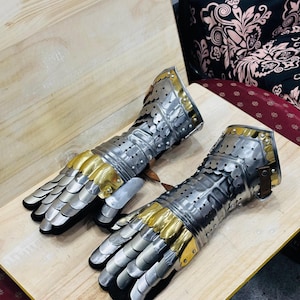 Puede incluir: Un par de guanteletes medievales, hechos de placas de metal plateado con detalles dorados. Los guanteletes están diseñados para proteger las manos y muñecas, con secciones de dedos articuladas y un forro negro. Se exhiben sobre una superficie de madera.