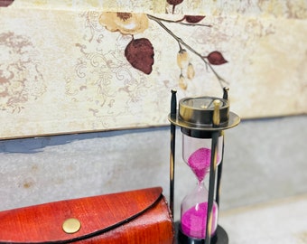 May include: A brass hourglass with pink sand and a leather case. The hourglass is about 5 inches tall and has a round base.
