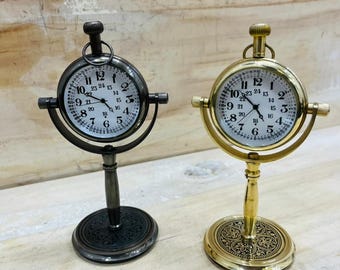 May include: Two small, decorative desk clocks. One is dark gray, the other is gold-toned. Both have a round clock face with black numerals and hands, and are mounted on a stand with a decorative base. The clocks are designed to resemble antique pocket watches.