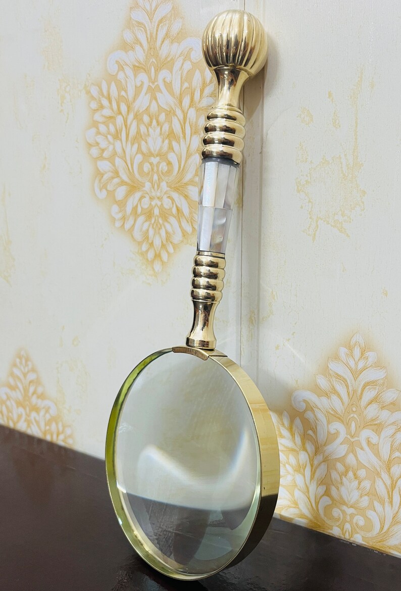 May include: A gold-coloured magnifying glass with a clear lens. The handle has a decorative design with a bulbous top, a section of white iridescent material, and a ribbed grip. A classic tool for reading and detailed work, it is a stylish accessory.