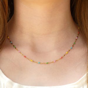 May include: A gold chain necklace featuring small, evenly spaced, multi-coloured enamel beads in shades of pink, green, blue, and yellow. The delicate metal links connect each bead, creating a continuous, vibrant, and lightweight circular design.