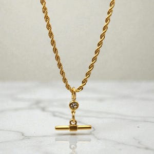 May include: A gold necklace with a twisted rope chain and a T-bar pendant. The pendant has a small, round, clear gemstone and a horizontal bar. The necklace is set against a marble background.