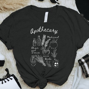 Puede incluir: Camiseta negra con un diseño blanco de temática de boticario. El diseño incluye ilustraciones de botellas, hierbas, un mortero y una maja, un caldero y una calavera. El texto en la camiseta dice "Apothecary, Elixir and Tonics, Medicinal, Herbs."