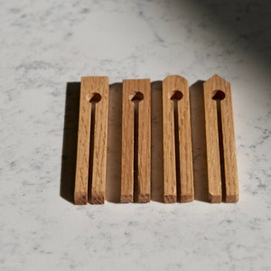 Puede incluir: Cinco pinzas de madera con la parte superior redondeada y un solo agujero en el centro de cada una. Las pinzas están dispuestas en fila sobre una superficie de mármol blanco.