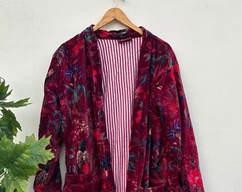 May include: A burgundy velvet robe with a floral pattern in red, green, and blue. The robe has a striped lining and two front pockets. It is hanging on a wooden hanger against a white wall.