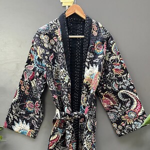 Op de afbeelding: Een zwarte, lange mouwen kimono met een kleurrijk bloemenpatroon. De kimono wordt in de taille vastgebonden met een bijpassende riem. De kimono is gemaakt van een gewatteerde stof en heeft witte stiksels.