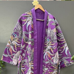 Op de afbeelding: Een paarse kimono-achtige badjas met een levendig bloemen- en vogelpatroon. De badjas heeft een effen paarse binnenkant en afwerking, met een bijpassende riem. Het ontwerp bevat witte, gele en blauwe accenten en is gemaakt van een gewatteerde stof.