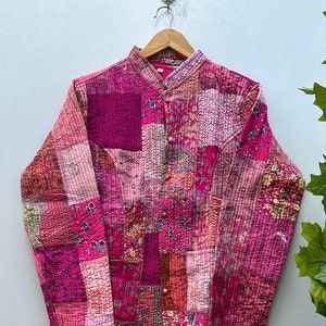 Peut inclure: Veste matelassée à patchwork dans des tons roses et magenta. Elle présente un col officier et des boutons. Le tissu est texturé, avec des coutures visibles. Le vêtement est suspendu à un cintre en bois, mettant en valeur son design.