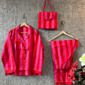 Könnte beinhalten: Ein passendes Pyjama-Set und eine Tragetasche mit rot-rosa Streifenmuster. Das Set beinhaltet ein Hemd mit Knöpfen, eine Hose und eine Tragetasche mit Klappenverschluss. Die Pyjamas hängen an Holzbügeln.