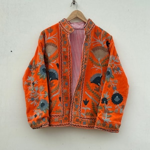 May include: An orange velvet jacket with detailed floral embroidery in blue, grey, and brown. The jacket has a mandarin collar and a red and white striped lining. It is displayed on a wooden hanger.