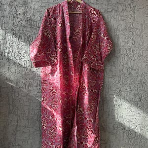 May include: A long, open-front robe with a floral pattern in shades of pink, red, and white. The robe is hanging on a wooden hanger against a textured gray wall. White trim outlines the edges and pockets.